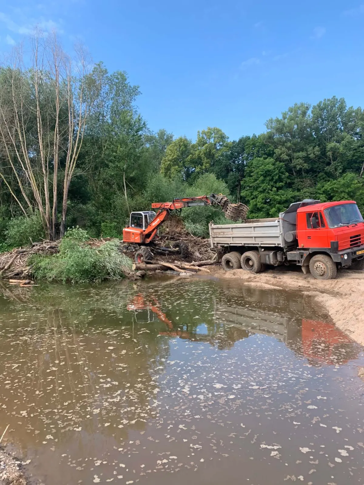 Červený bagr s drapákem nakládá materiál z břehu na nákladní vůz