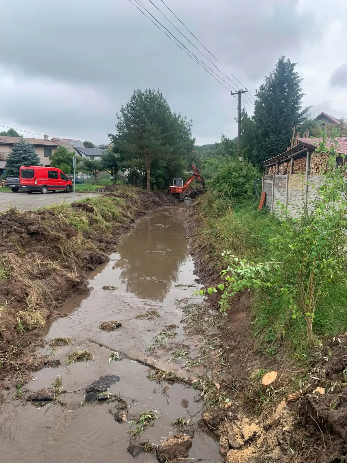 Vyčištěné koryto potoka vede podél silnice mezi domy, v dálce bagr